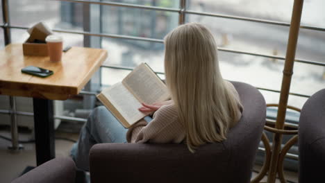 back view of student preparing for exam, focused as she flips to a new page of her textbook while sitting at a table in a study area with a cup of coffee and phone beside her