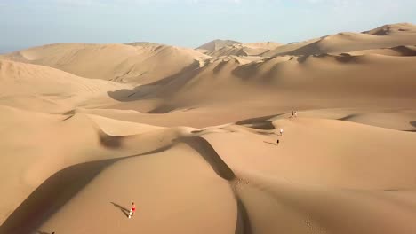 Aerial,-tracking,-drone-shot,-of-people-and-buggy-vehicles,-on-sand-dunes,-near-the-Huacachina-oasis-village,-on-a-sunny-day,-in-southwest-Peru