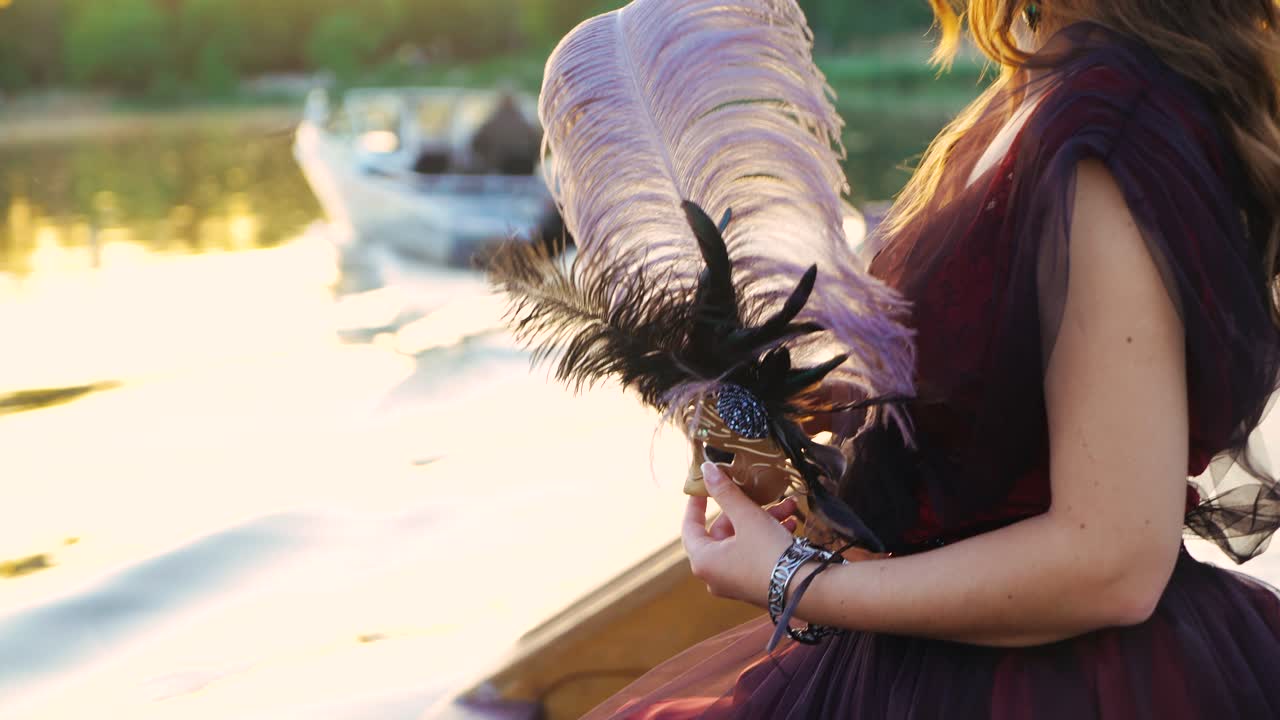 Luxury woman removing carnival mask from her face and looking at powerboat floating nearby at sunset. Beautiful lady in dress holding mask on face standing over the water. Close-up