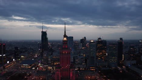 Volar-Hacia-Atrás-Sobre-La-Ciudad-De-La-Noche.-Vista-Panorámica-Aérea-De-Los-Edificios-Del-Centro-De-Gran-Altura-Al-Atardecer.-Varsovia,-Polonia