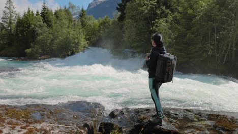 female traveler with a backpack, drinking water in nature in the forest near a mountain river.