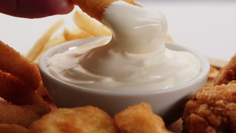 hand dipping fried chicken nuggets and onion rings into sauce with french fries