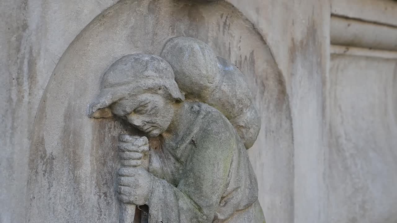 Visuale ravvicinata di una figura commemorativa in pietra usurata, appoggiata a un bastone, che cattura la quieta emozione del ricordo all'interno del cimitero di Bunhill Fields