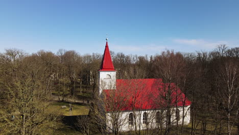 Vista-Aérea-De-La-Iglesia-Luterana-De-Lielvarde-En-La-Orilla-Del-Río-Daugava,-Iglesia-Blanca-Con-Techo-Rojo-A-Través-De-Los-árboles-Sin-Hojas,-Día-Soleado-De-Primavera,-Tiro-Ancho-Avanzando