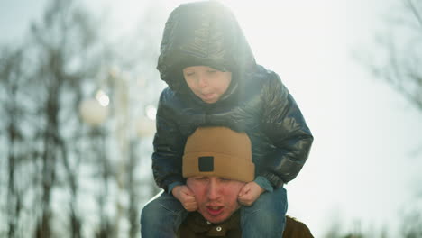 padre llevando a su hijo sonriente en sus hombros, mientras el niño juega con su barbilla, haciendo que el rostro del padre se gire, rojo por el peso del niño, con suaves rayos de luz por detrás