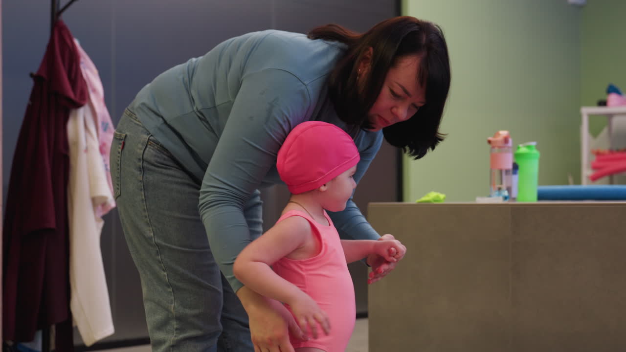 Caregiver carrying girl child down in front of pool, water bottle on table by side, secure hold, gentle guidance, bright daylight on blue water, summer mood, calm scene, safe approach to swim zone