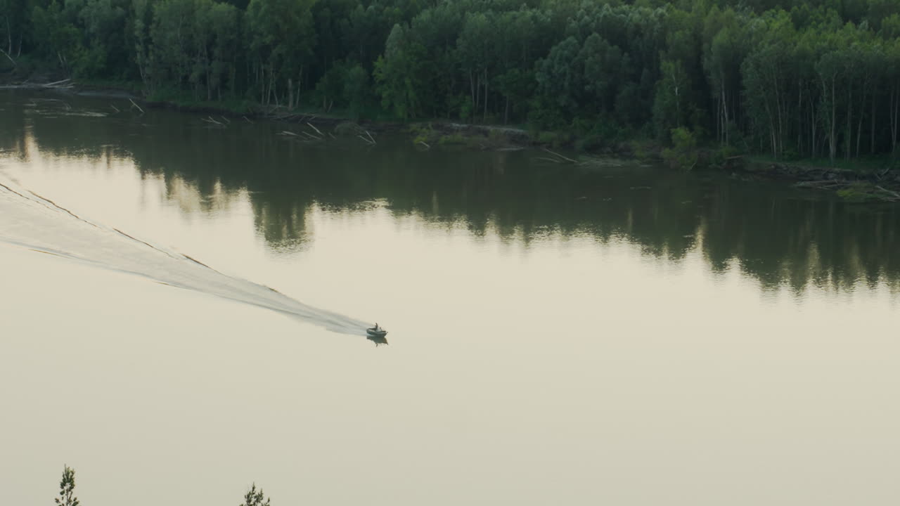 small boat cuts across calm river surface leaving ripples behind as it moves toward forested riverbank lined with dense trees under soft evening light in peaceful and remote natural setting