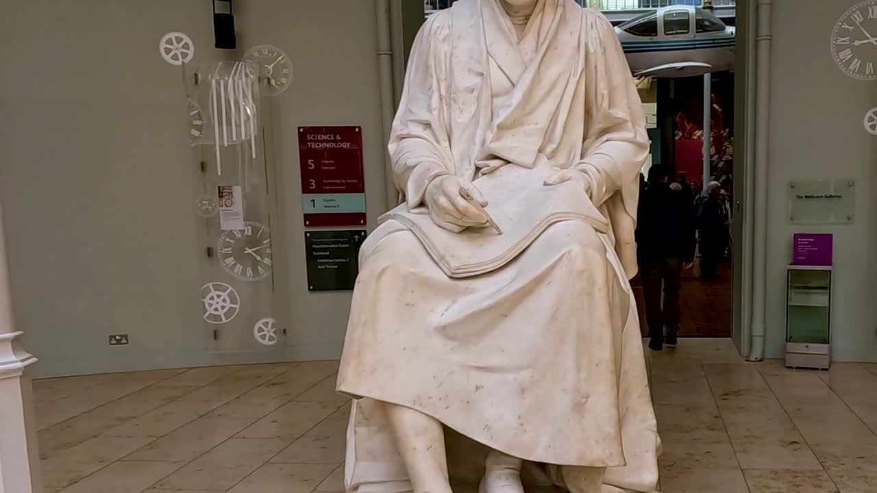 A close-up of a seated marble statue with visible signage and museum visitors in the background.