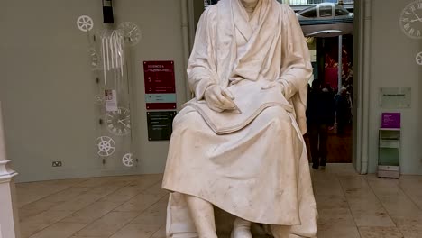 a close-up of a seated marble statue with visible signage and museum visitors in the background.