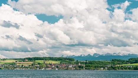 towering cumulus clouds dominate the sky over the charming lakeside town with alpine peaks in the distance (lake murten, fribourg, switzerland). the glistening lake creates a breathtaking timelapse.
