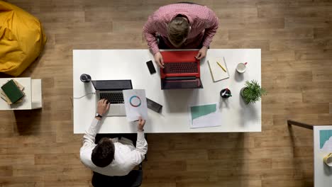 young men working on laptop, giving document to colleague, topshot, sitting at table in modern office, work concept