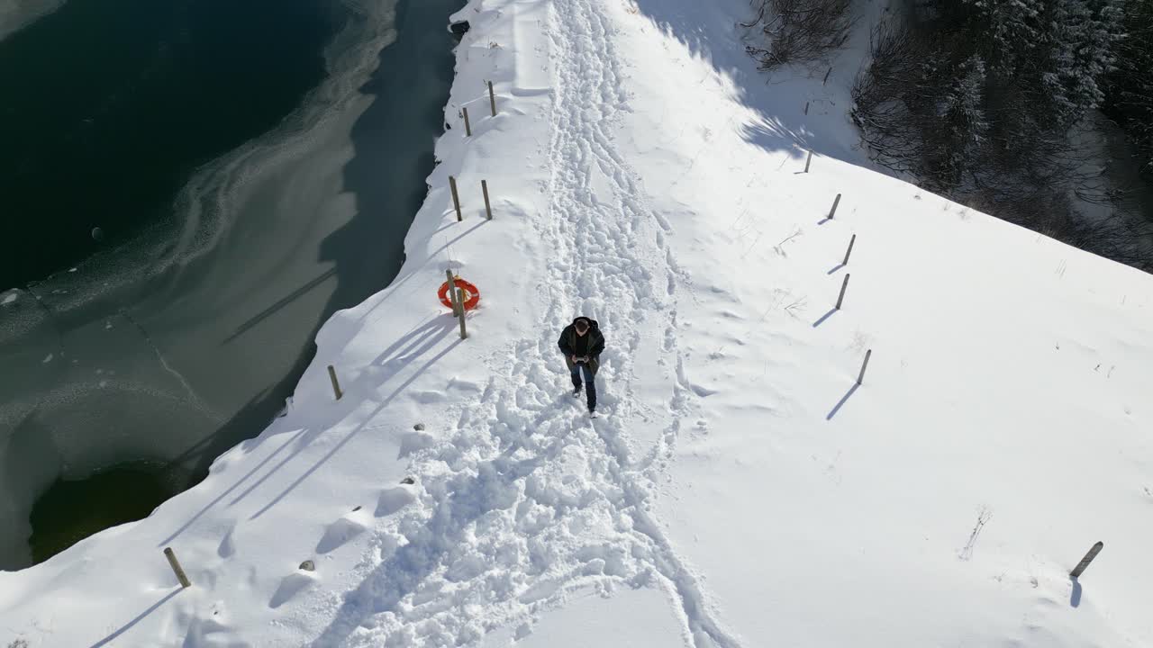 con la cabeza inclinada, un hombre está caminando por un camino nevado al lado del lago congelado, ubicado en la cima de una montaña en engelberg, brunni, en bahnen, suiza