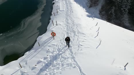 with head bowed down, a man is walking on a snowy path beside the frozen lake, located on top of a mountain at engelberg, brunni, in bahnen, switzerland