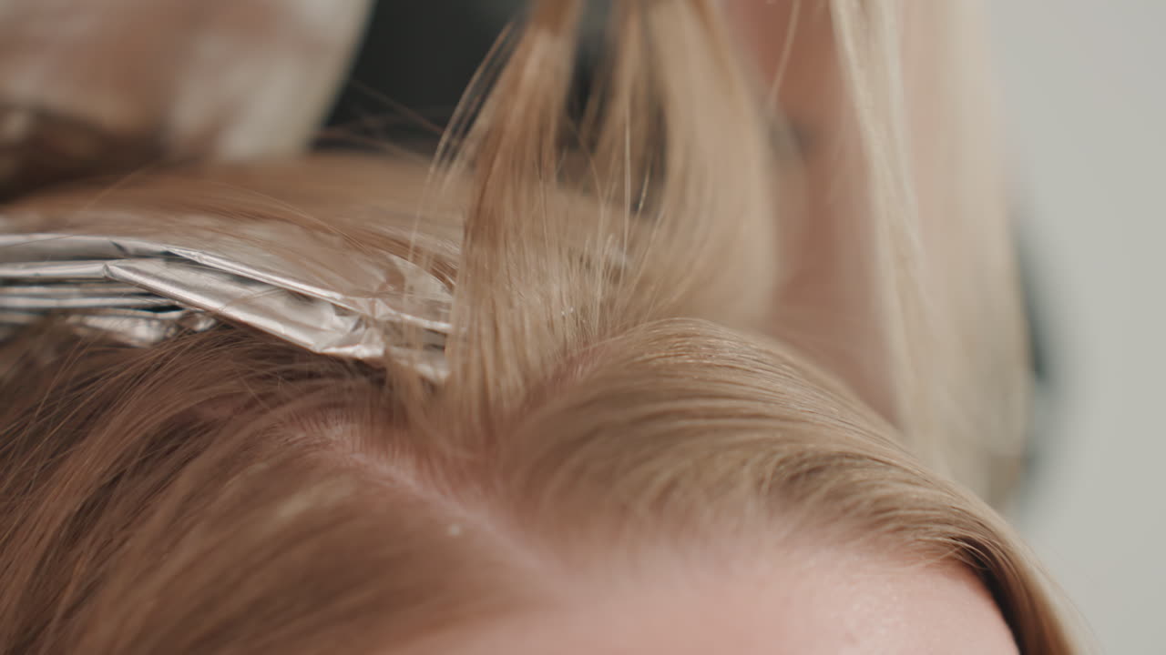 Primo piano di una donna caucasica che fa dei colpi di luce ai capelli con la stagnola in un salone; la stilista separa delle ciocche bionde sottili, applica il decolorante con un pennello, piega la stagnola sulle radici, liscia le ciocche vicino al cuoio capelluto, studio neutro