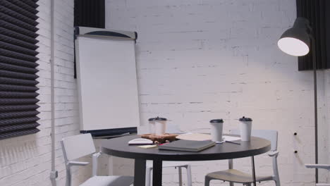 empty small meeting room with round black table, white chairs, takeaway coffee, notebooks, floor lamp and whiteboard