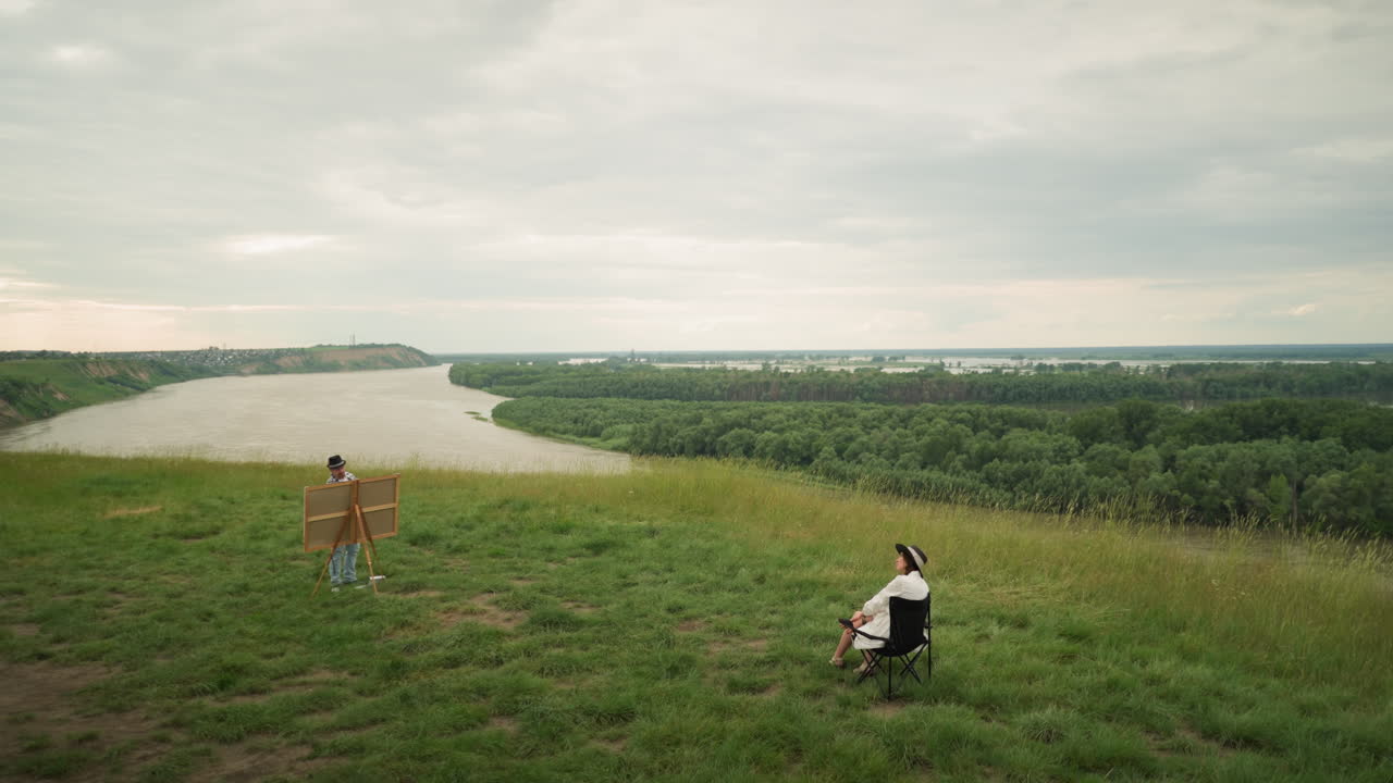 A woman in a hat, sitting comfortably on a chair in a grassy field, while an artist paints her near a serene lake. The expansive landscape, with its peaceful atmosphere and cloudy sky
