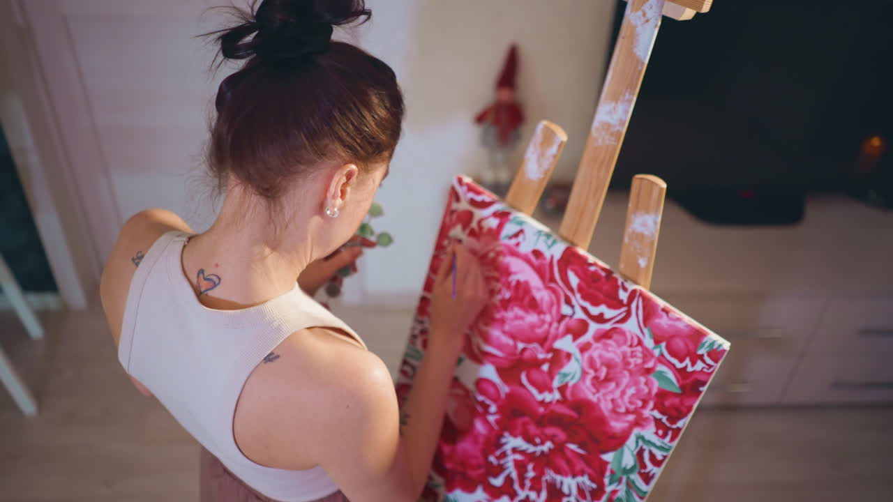 Overhead White Woman Painting Floral Canvas At Easel, Studio Rehearsal Vibe, Back View With Bun And Small Neck Tattoo, Scattered Supplies, Dynamic Brush Movement, Warm Home Studio Scene