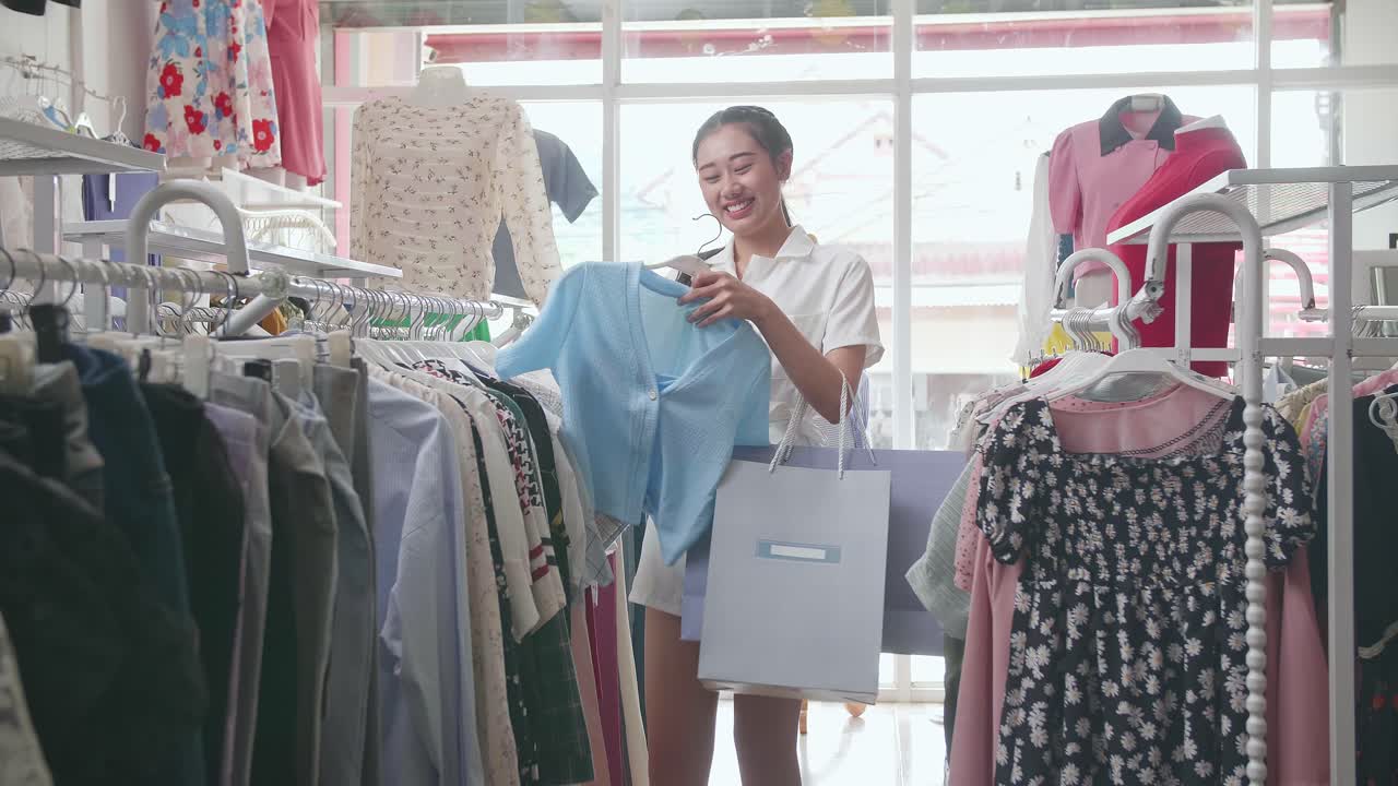 쇼핑 가방을 들고 있는 젊은 아시아 여성이 파란색 옷을 선택하고 미소 짓고 엄지손가락을 보여주며 부티크에서 쇼핑하는 동안 카메라에 몸짓을 합니다.