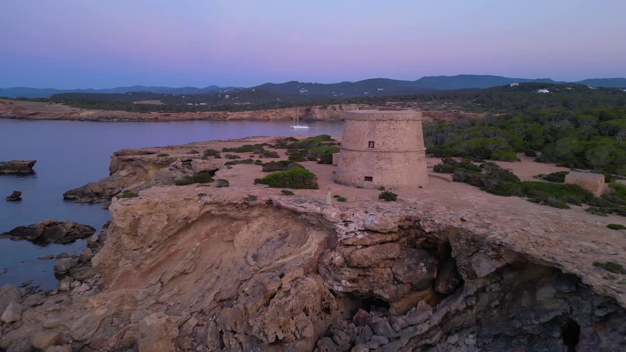 Barefoot hippie girl practicing yoga at sunset near Torre des Carregador, Ibiza, Spain. Amazing aerial view flight fly reverse drone