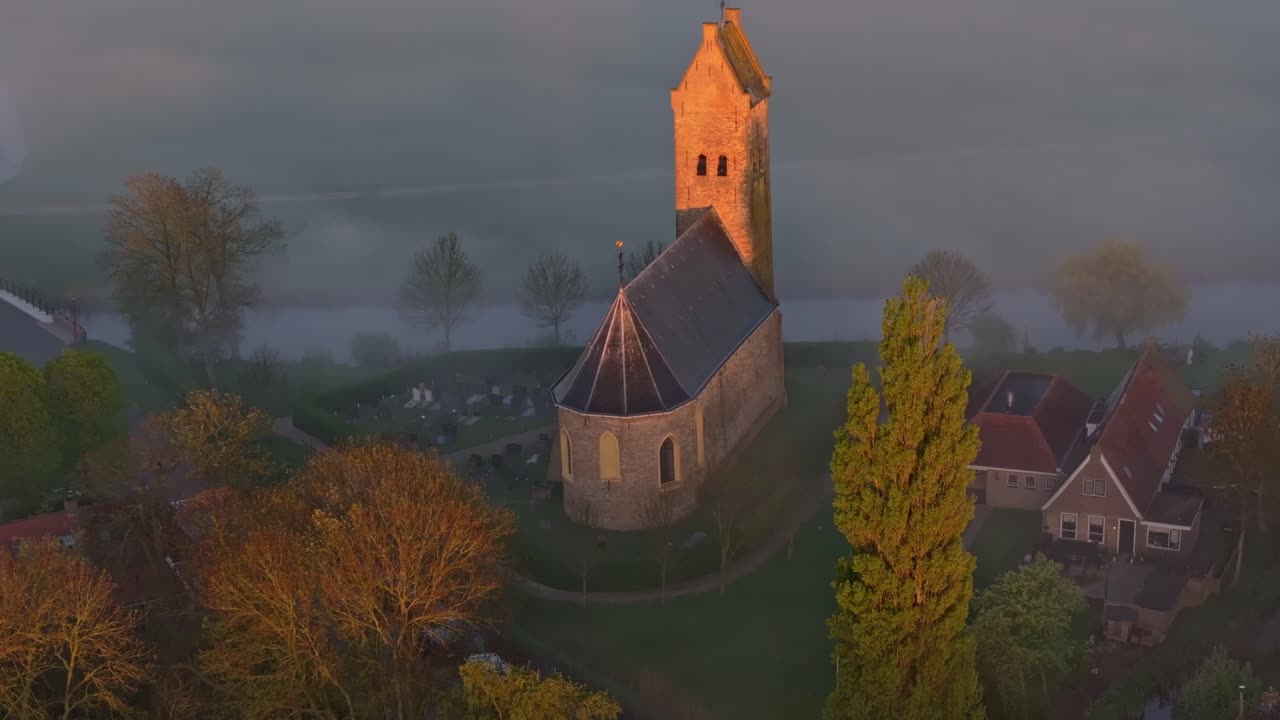 Luftaufnahme eines dunstigen Sonnenaufgangs hinter Dorfkirche und Wasser, Friesland
