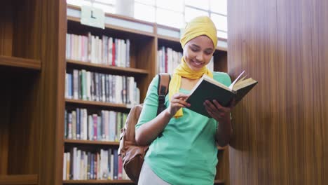 una estudiante asiática con un hijab amarillo apoyada en estantes de libros y leyendo un libro
