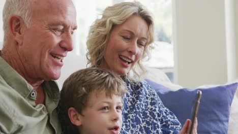 Glückliche-Kaukasische-Großeltern-Mit-Enkel,-Der-Tablet-Benutzt-Und-Im-Wohnzimmer-Sitzt