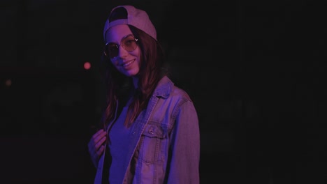 adolescente caucásica con gafas y gorra sonriendo y posando para la cámara al aire libre por la noche