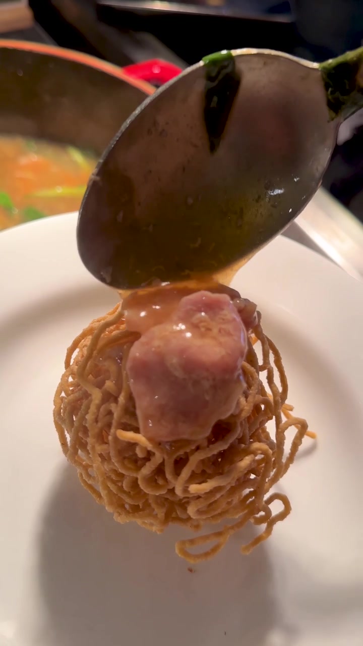 Un cucchiaio versa del sugo denso di maiale su una palla di noodles croccanti in un piatto.