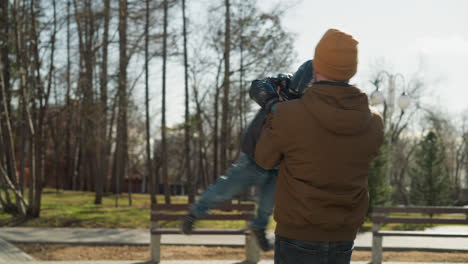 a close-up of a father and son enjoying outdoor playtime as the father spins his son gently in a park, with trees and benches in the background