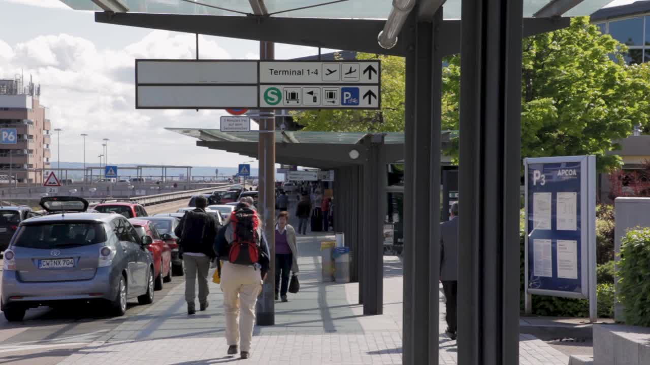 Travelers walk past cars and signage outside a busy airport terminal
