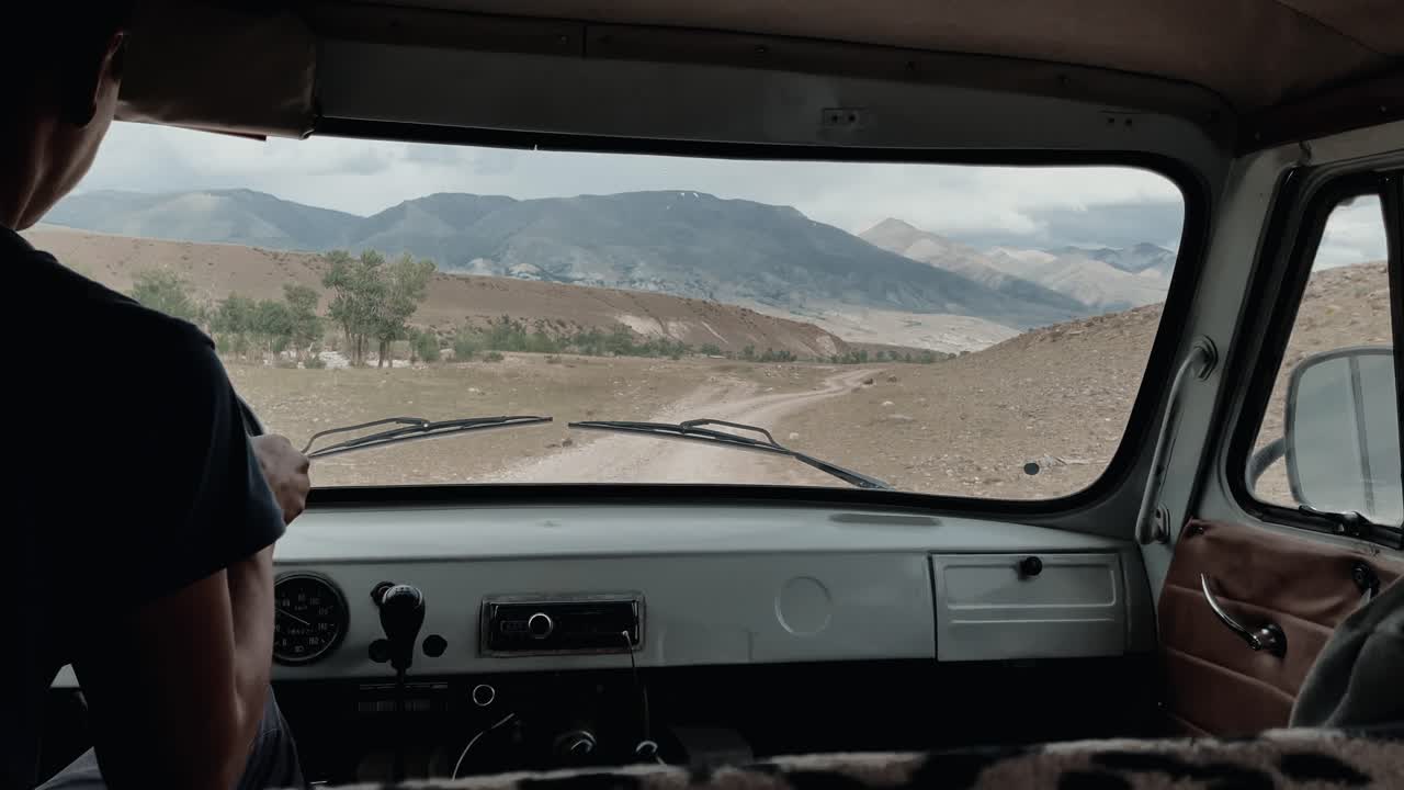 Blick aus dem Inneren eines Geländewagens, der auf einer Schotterstraße durch das Altai-Gebirge, Russland, fährt, mit weiter Tallandschaft, Hügeln und bewölktem Himmel voraus.