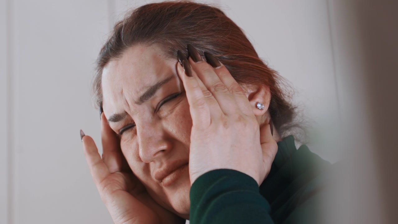 Mulher branca segurando as têmporas com dor em casa, close-up apertado mostrando rosto avermelhado e expressão tensa, dedos pressionando as têmporas, unhas curtas, suéter verde, atmosfera íntima transmitindo enxaqueca.