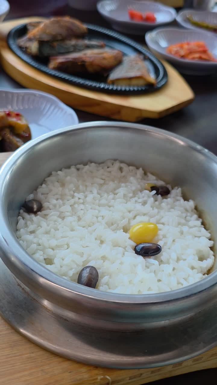 Vertical parallax close-up of a steaming bowl of Korean nutritious rice with beans and ginkgo, with a sizzling plate of grilled fish and side dishes in the background