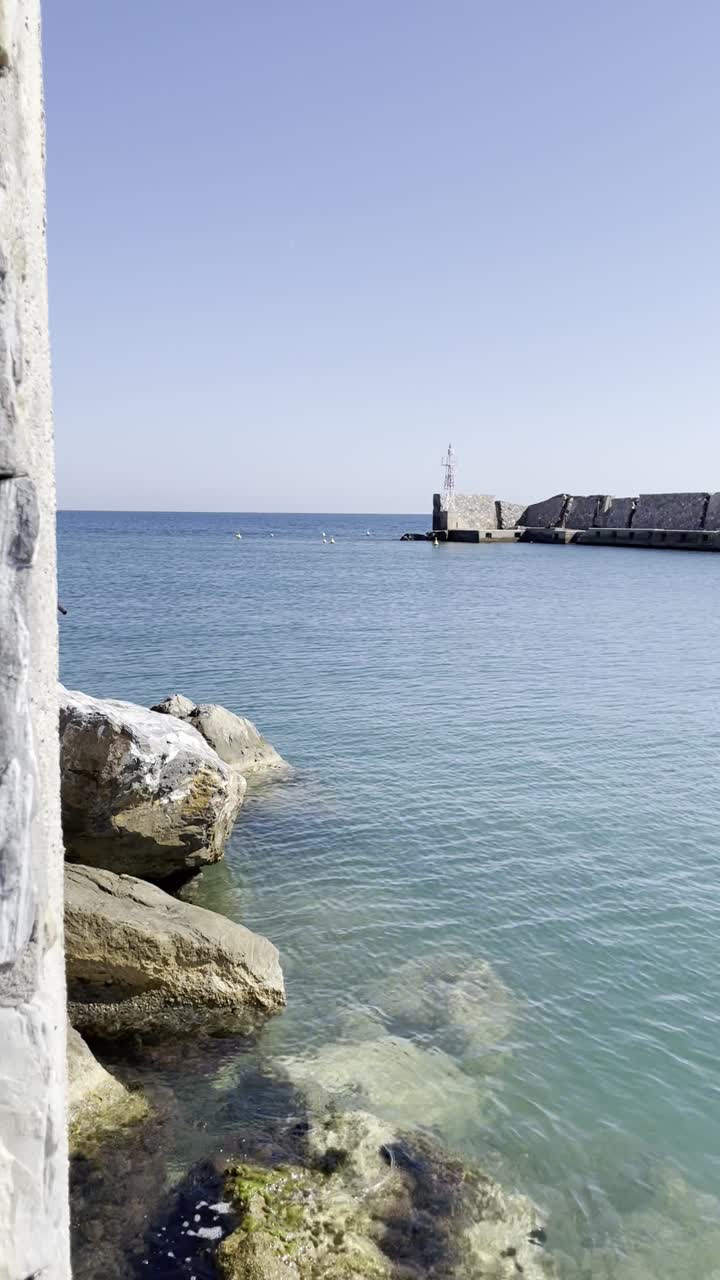 A tranquil scene of a stone breakwater extending into clear blue waters, framed by rocky shores and a bright sky, with birds dotting the horizon