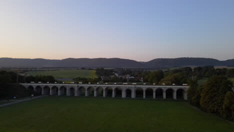 Vista-Aérea-De-Un-Viaducto-De-Tren-Con-El-Campo-Circundante-Y-La-Ciudad-Al-Fondo