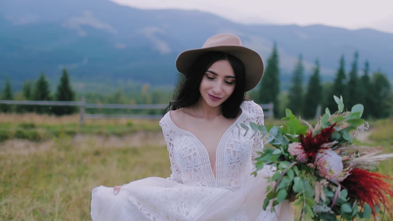 Portrait of lovely dark-haired bride in nature. Beautiful woman in stylish wedding dress and a hat posing on camera in the mountainous area.