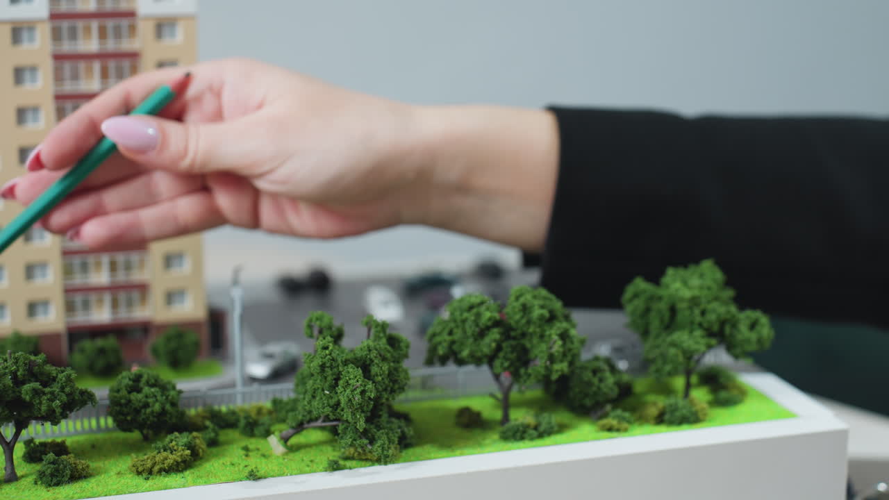 Close up of professional hand using pencil to point at tree on miniature urban model during project discussion, with car key nearby on office desk, suggesting planning