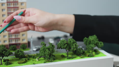 close up of professional hand using pencil to point at tree on miniature urban model during project discussion, with car key nearby on office desk, suggesting planning