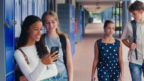 two female high school or secondary students looking at social media or internet on phone by lockers