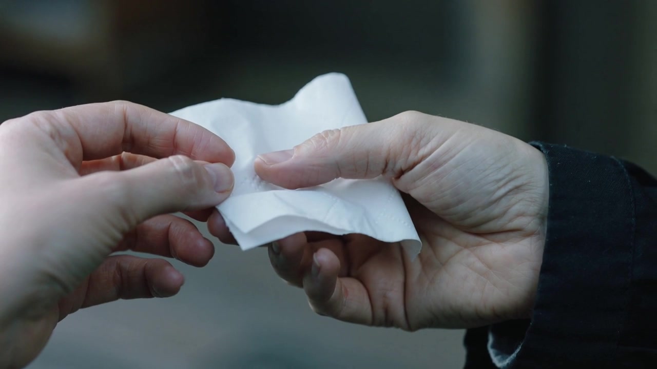 Mani adulte che si incontrano passandosi un biglietto accartocciato sul marciapiede, la mano destra con un polsino scuro, la sinistra che si estende.