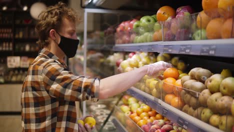 giovane in camicia a quadri, con maschera nera e guanti di plastica trasparenti, fa la spesa per la frutta, sceglie i limoni al supermercato durante la pandemia di quarantena del coronavirus covid-19. vista laterale