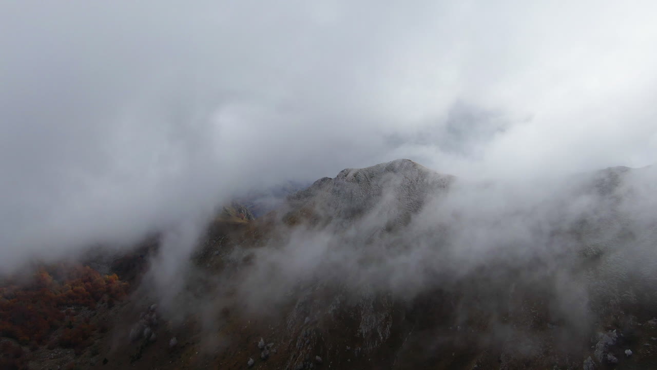 The cold, misty fog blankets the mountain top, slowly rolling over it as the drone flies above on an autumn morning