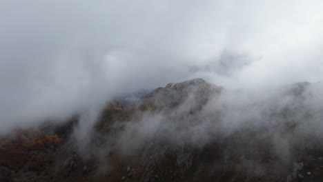 the cold, misty fog blankets the mountain top, slowly rolling over it as the drone flies above on an autumn morning