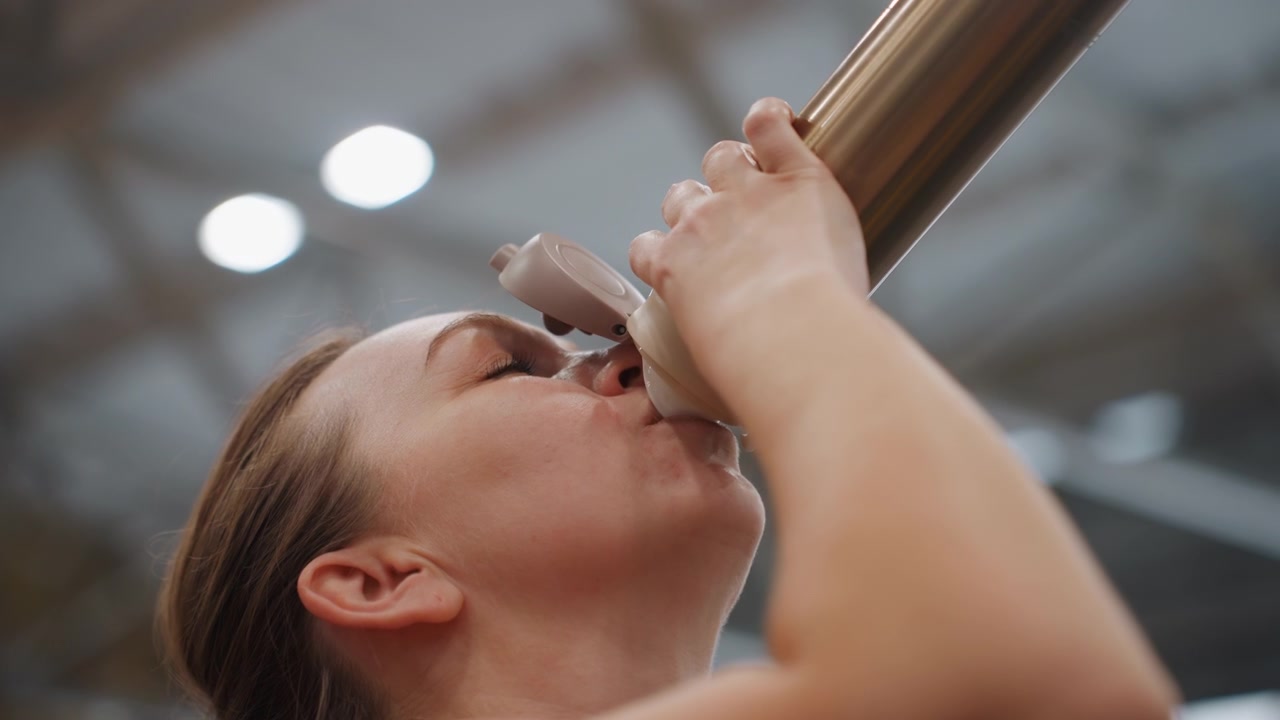 Primo piano di una donna bianca dopo una partita di badminton che sorseggia una bevanda, mostrando sollievo e soddisfazione, con sudore sulla fronte, mentre tiene un thermos dopo la partita, con le luci dell'arena e una folla sfocata sullo sfondo, in un momento tranquillo di recupero e calma.