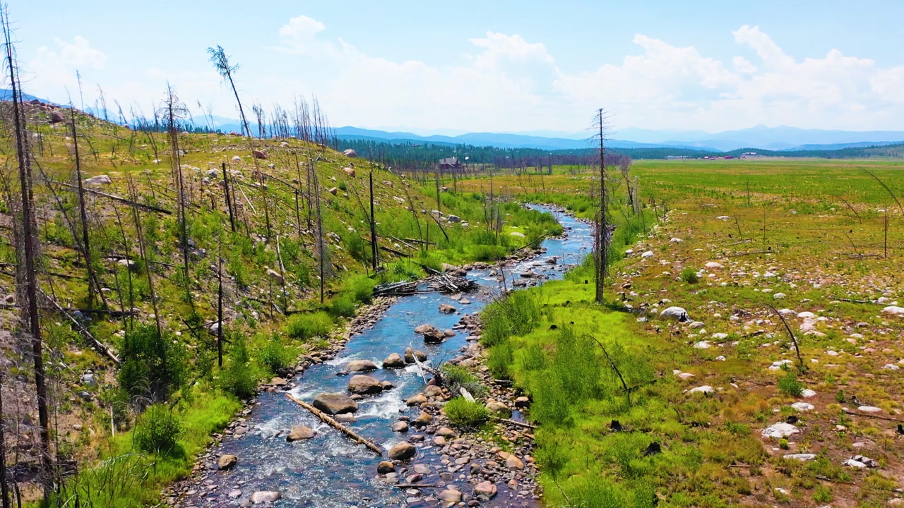 Cinematic aerial retreating above a Montana style river through wide western meadowland with stones green creek margins burned trees and long mountain vistas beneath bright clouds