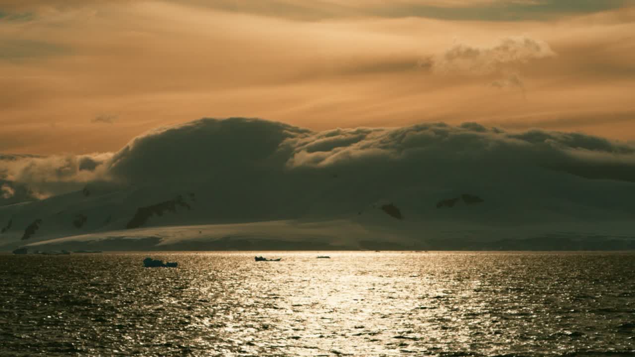 Epischer, filmischer Sonnenuntergang in der Antarktis mit goldener Meeresreflexion, bedrohlichen dunklen Sturmwolken und silhouettierten Zodiac-Booten am Horizont.