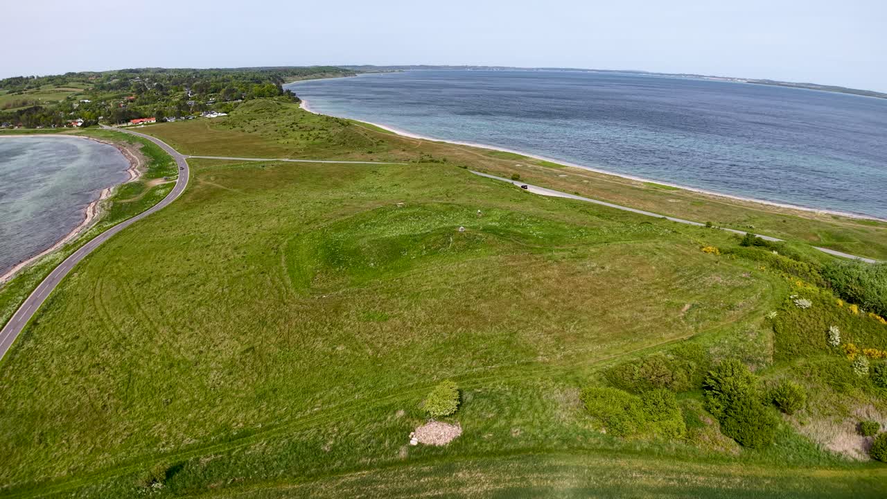 Drone shot of a narrow coastal peninsula with grassy meadows and a dirt path between two bodies of water. Footage pans along the shoreline, showing the green headland, blue sea and distant islands