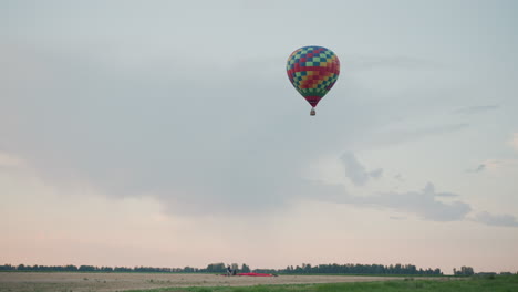 colorful checkered hot air balloon carrying passenger ascending slowly over vast open field with vivid burner flame lighting wicker basket against soft pastel sky at sunset