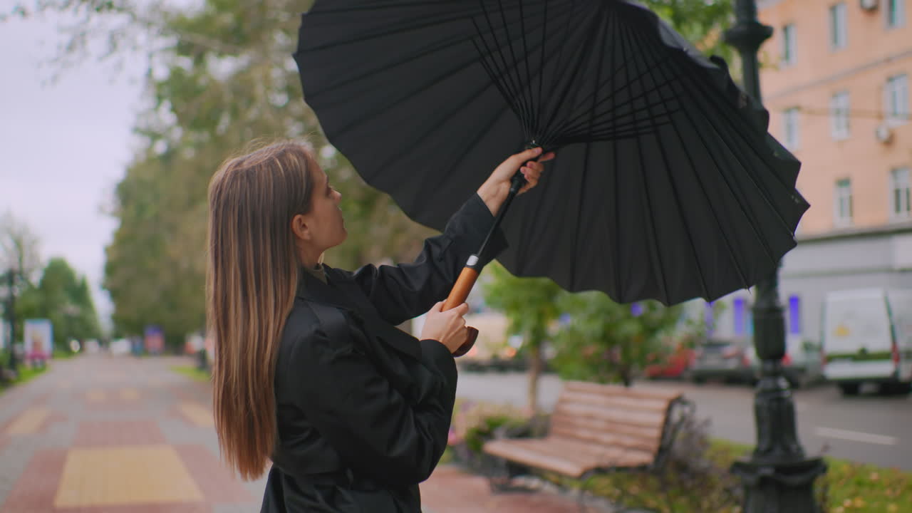 Amazing lady in black coat closing umbrella on rainy city street with trees bench and buildings in background preparing for rain protection outdoors urban lifestyle weather season change concept