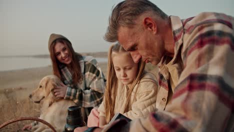 Close-up-shot-of-a-happy-brunette-man-with-gray-hair-in-a-checkered-shirt-showing-his-little-daughter-a-book-and-telling-him-what-it’s-about,-his-wife-and-a-big-dog-are-near-them-during-a-picnic-in-the-summer-outside-the-city